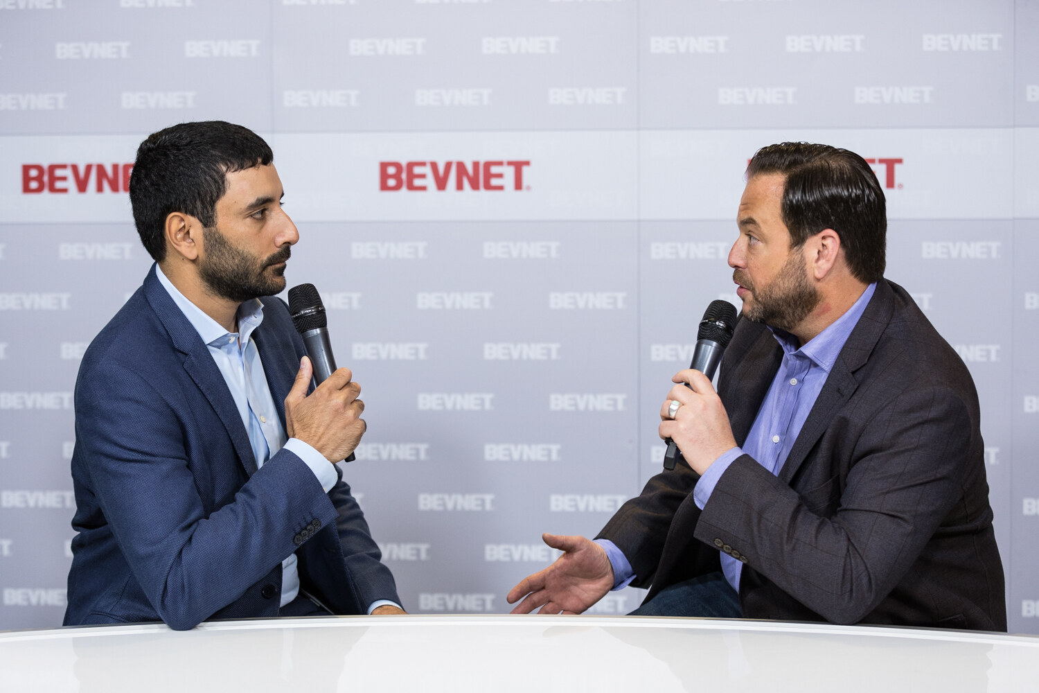 Two men with microphones talk across a white desk in front of a BevNET-patterned wall.