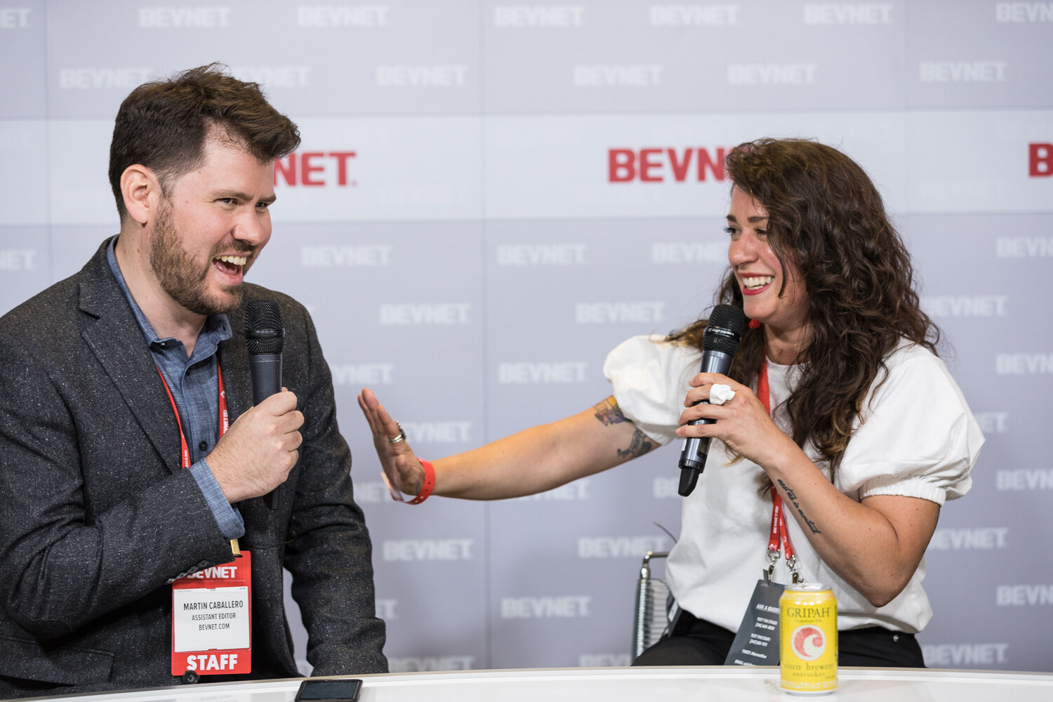 A bearded man with a staff badge and a woman in a white puff-sleeve top laugh together at the Livestream Studio desk, with a canned drink in front of her.