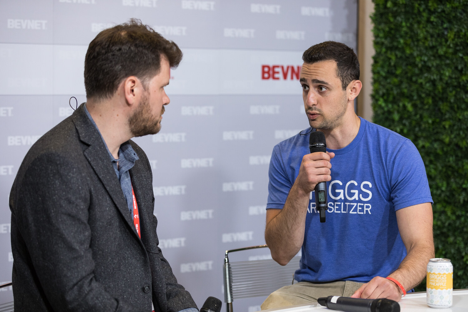A man in a blue t-shirt that mentions seltzer speaks into a microphone while another man listens, with a can on the table.