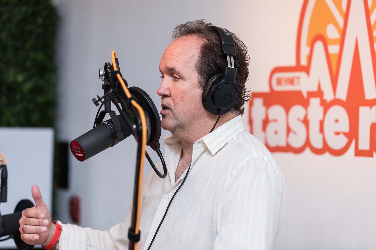 A man in a cream shirt and headphones gestures toward a studio microphone in front of a Taste Radio logo wall.