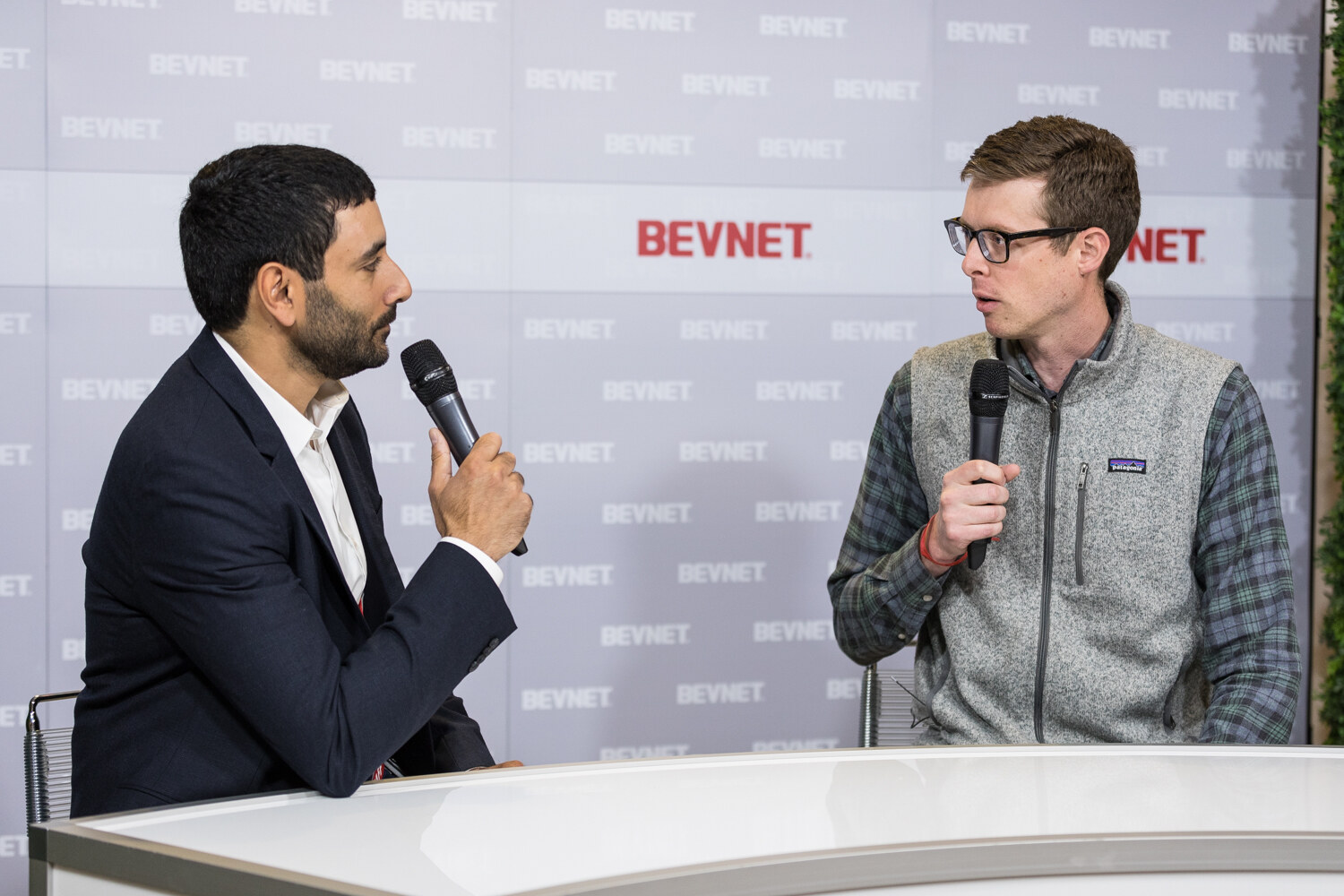 A man in a navy blazer speaks into a microphone while a man in glasses and a gray Patagonia vest answers.
