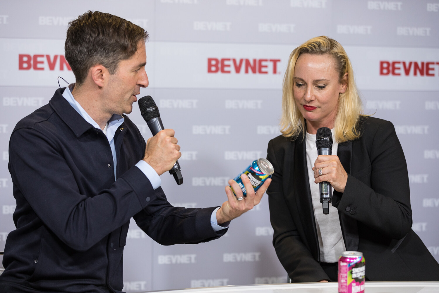 A man in a dark jacket holds a can toward a blonde woman in a black blazer, both with microphones, at the Livestream Studio desk.