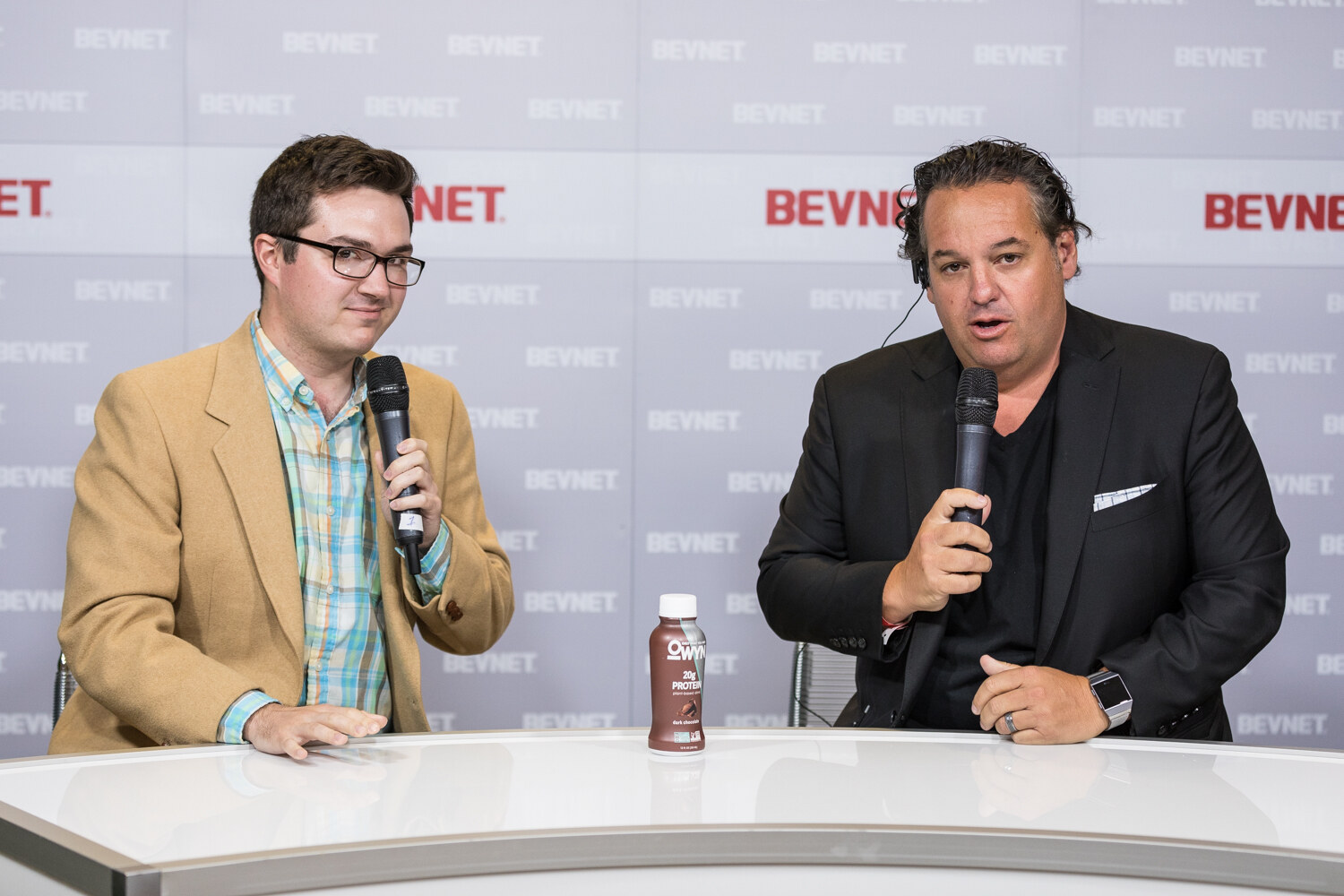 A man in a tan jacket and a man in a black jacket with a pocket square, both holding microphones, sit at a white desk.