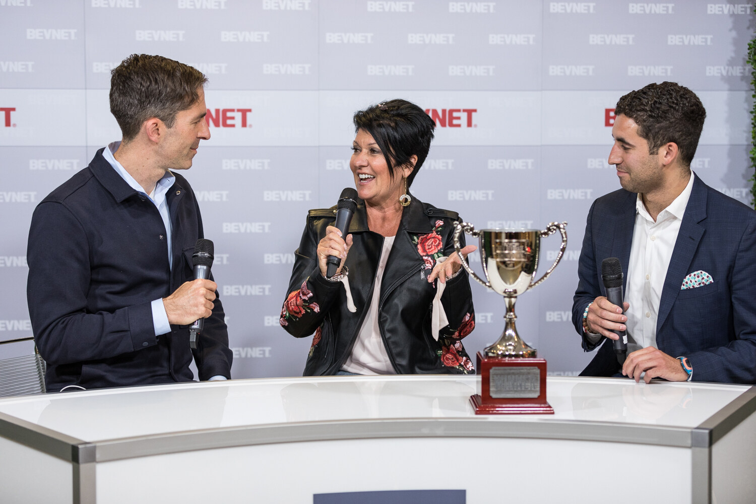 A man in a navy jacket, a woman in a black floral jacket and another man in a navy blazer sit at a white desk beside a silver trophy.