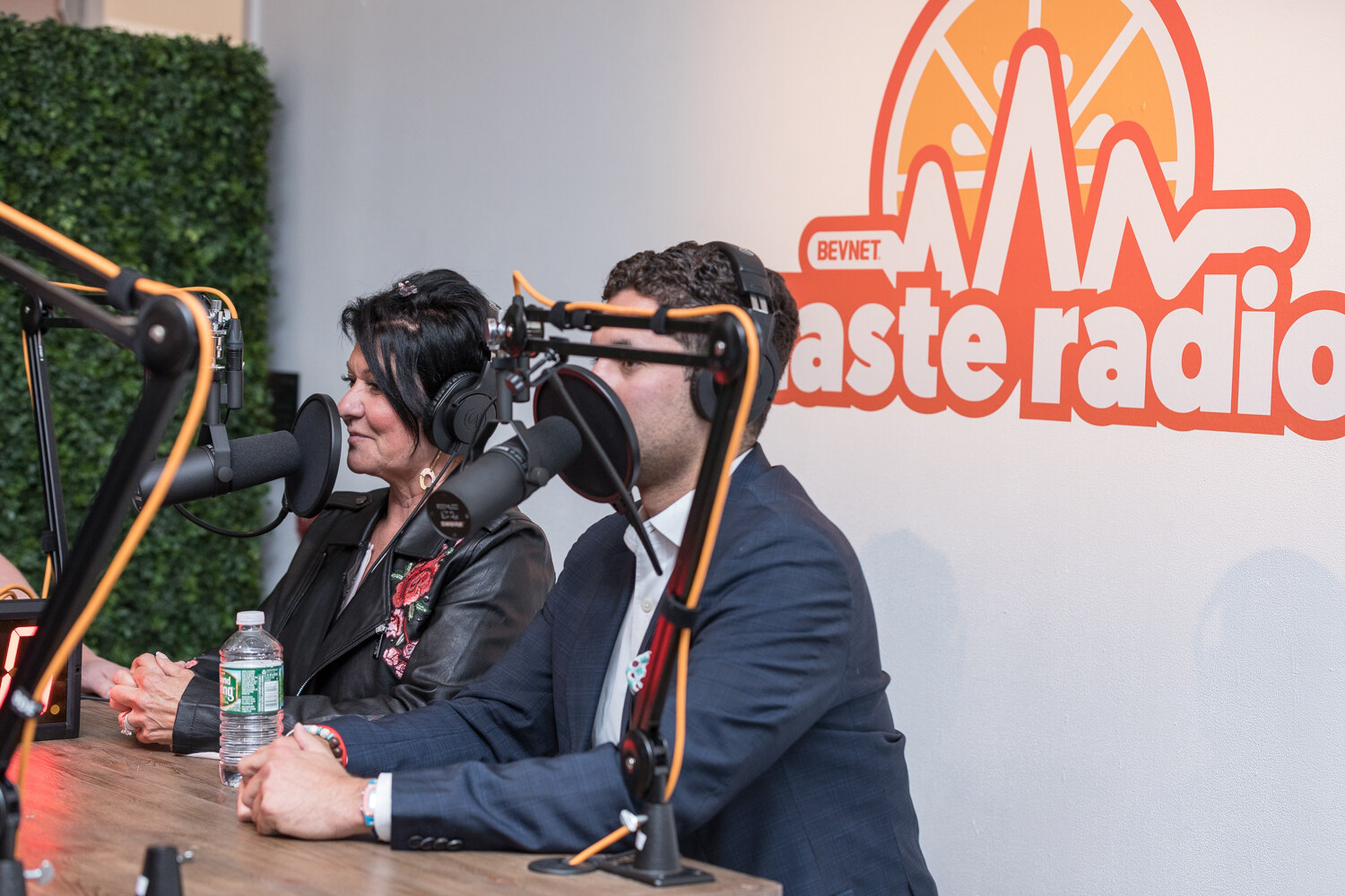 A woman in a black floral jacket and a man in a navy blazer sit at microphones in front of a Taste Radio logo.