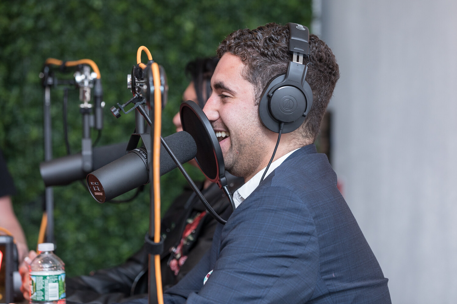 Side view of a smiling man in headphones in front of a studio microphone with a hedge wall behind him.