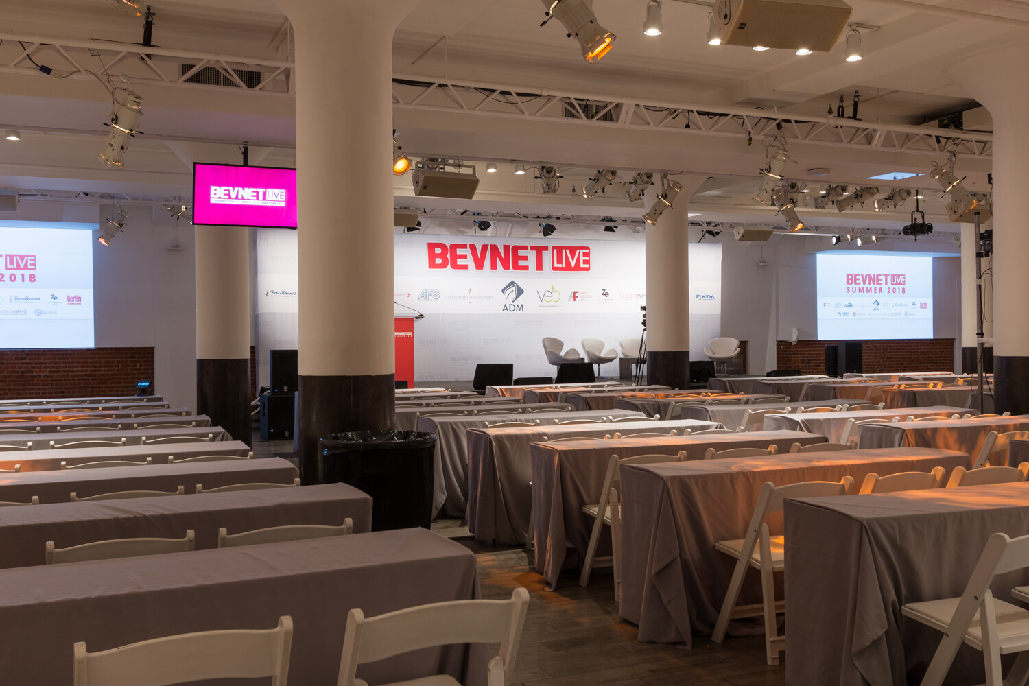 Rows of draped tables with chairs facing a stage with BevNET Live signage, columns and screens.