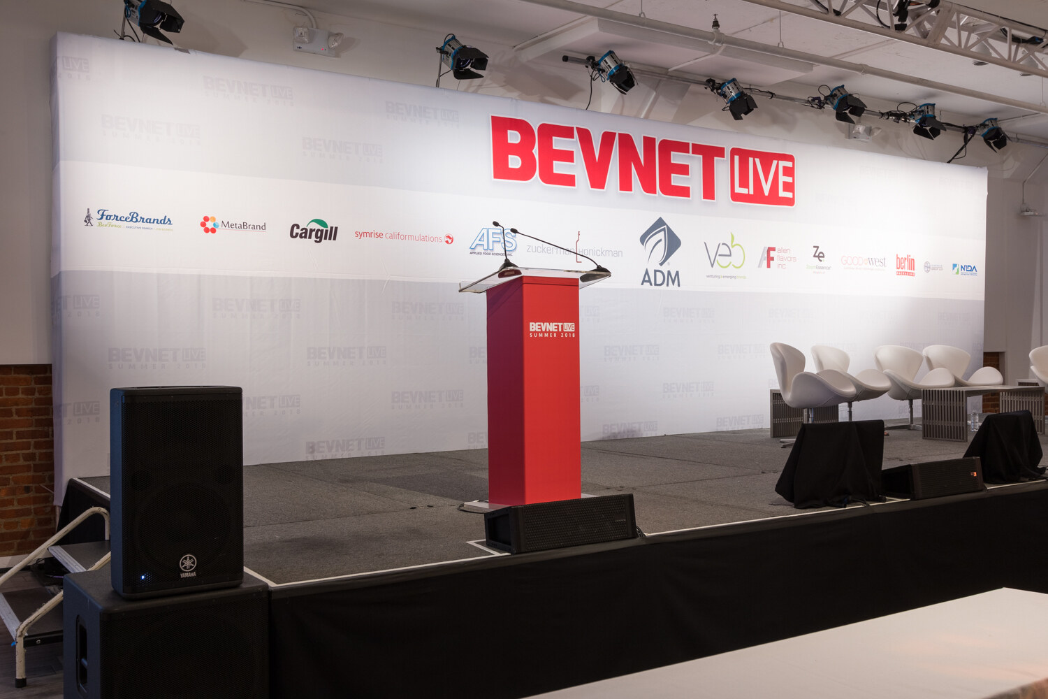 A long stage with a BevNET Live backdrop, a red lectern, speakers and white swan chairs.