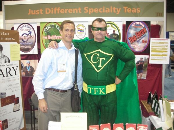 A man in a light blue shirt stands beside a person in a green superhero costume with a mask and cape at the Just Different Specialty Teas booth, with Tea For Men and Tea Force posters behind.