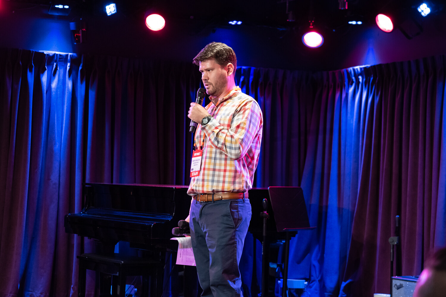 Presenter in an orange plaid shirt with a STAFF badge holding a microphone on the club stage beside a piano.
