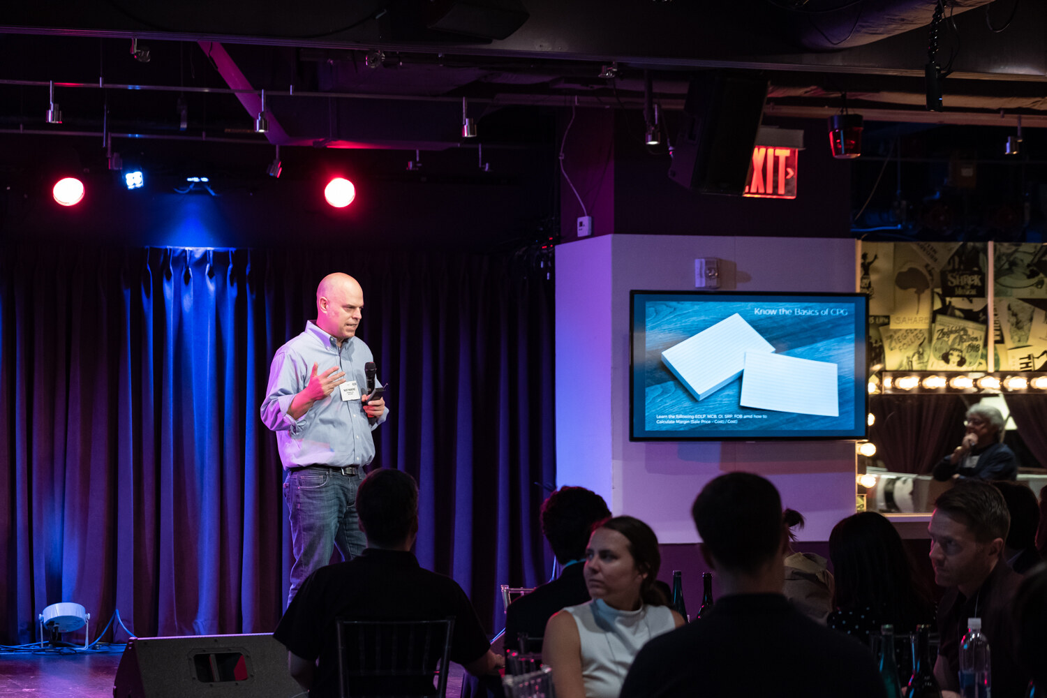 Presenter in a light blue shirt on stage beside a wall monitor with a slide while attendees watch from tables.