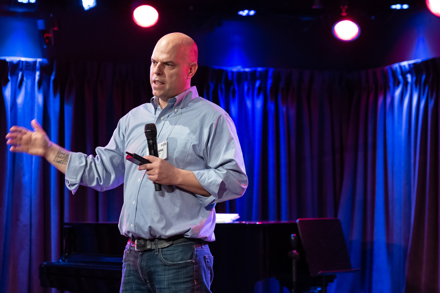 Presenter in a light blue shirt reaching out with one arm while speaking under red and blue stage lights.