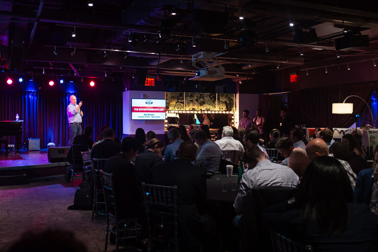 Wide view of the Beverage School room with a presenter on the club stage and attendees at round tables.