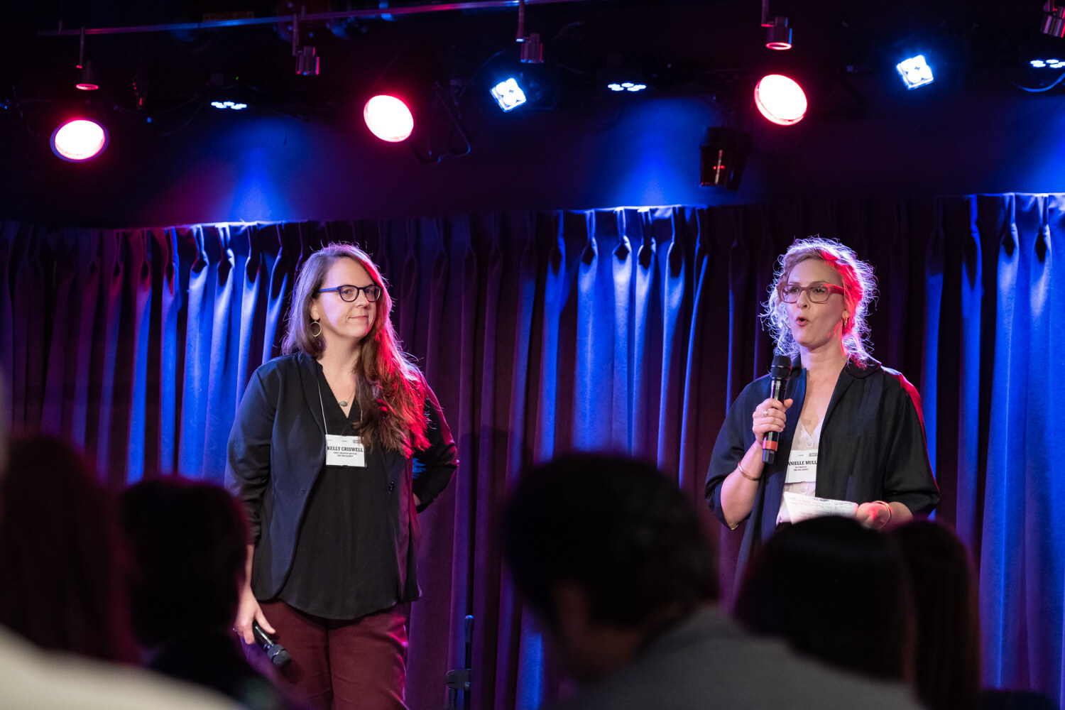 Two women presenters in dark cardigans on the club stage under red and blue stage lights, one holding papers.