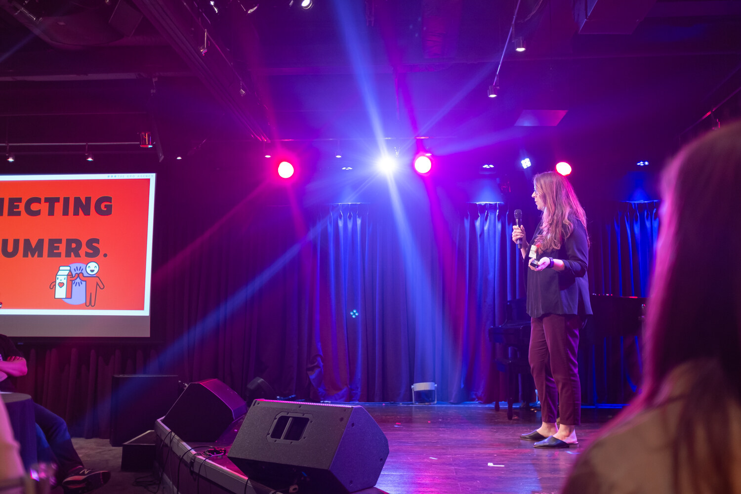 Woman presenter in a dark blazer standing on stage beside a red slide that reads Connecting Consumers.