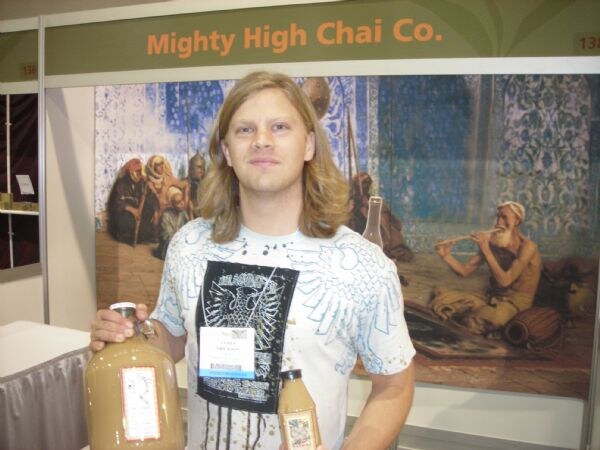 A man with shoulder-length hair and a graphic t-shirt holds two bottles of chai in front of a painting at the Mighty High Chai Co. booth.