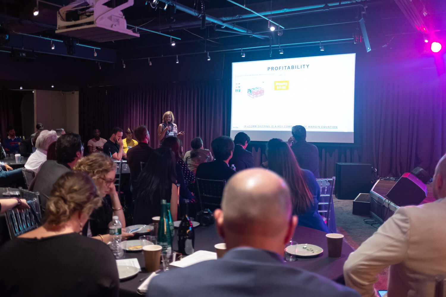 Attendees at round tables facing a screen titled Profitability while a presenter stands at the side.