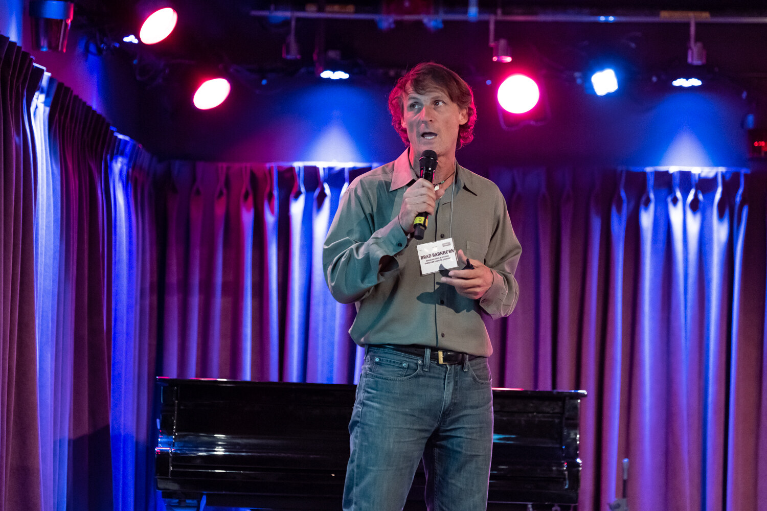Presenter in an olive button-up shirt and name badge speaking into a microphone in front of purple curtains and a piano.