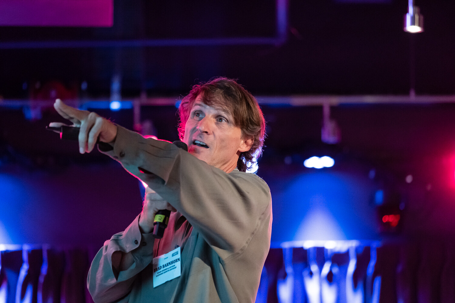 Close view of a presenter in an olive shirt pointing up and out toward the audience.