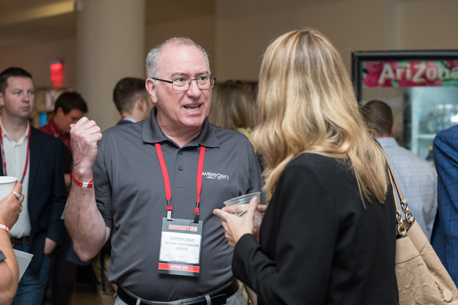 Man in a gray polo and lanyard talking animatedly with a woman with long blond hair.
