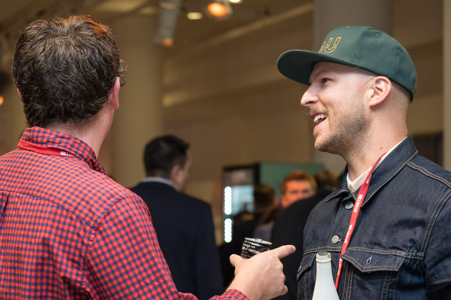 Attendee in a denim jacket and green cap laughing while talking with another attendee.