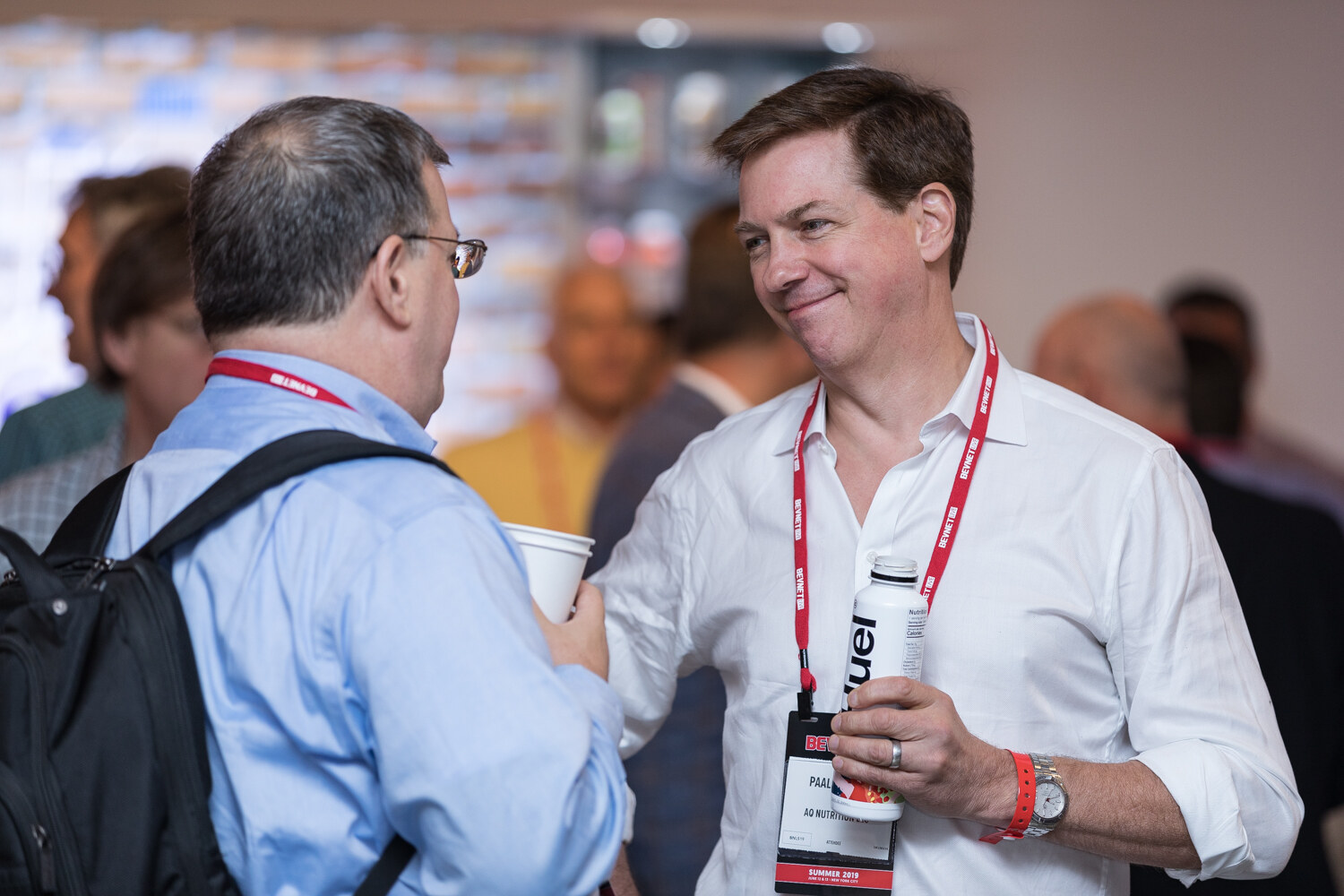 Smiling attendee in a white shirt holding a cup and a bottle while talking with another guest.