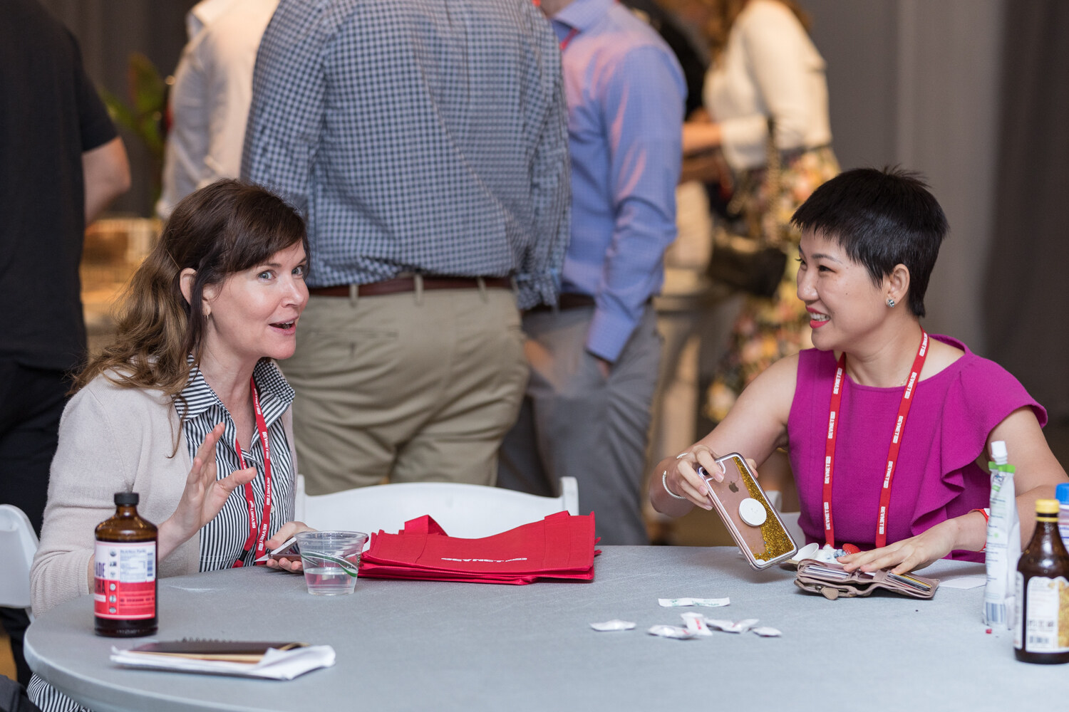 Two women seated at a round table talking with samples and red tote bags in front of them.