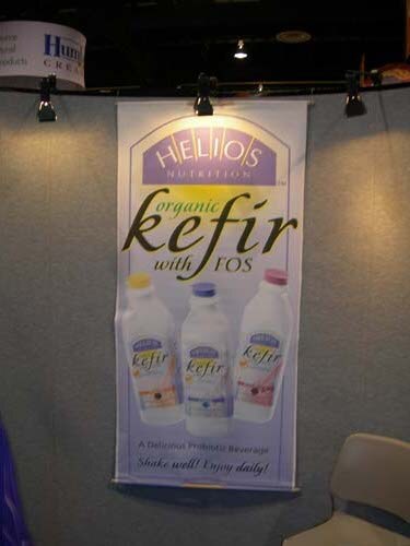 Hanging banner reading Helios Nutrition organic kefir with FOS, showing three bottles of kefir, lit by track spotlights on a gray wall; a chair is at the lower right.