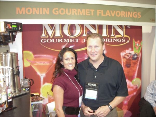 A woman in a maroon sweater and a man in a black polo stand together at the Monin Gourmet Flavorings booth, with a Bunn brewer and syrup bottles at left and cocktail graphics behind.