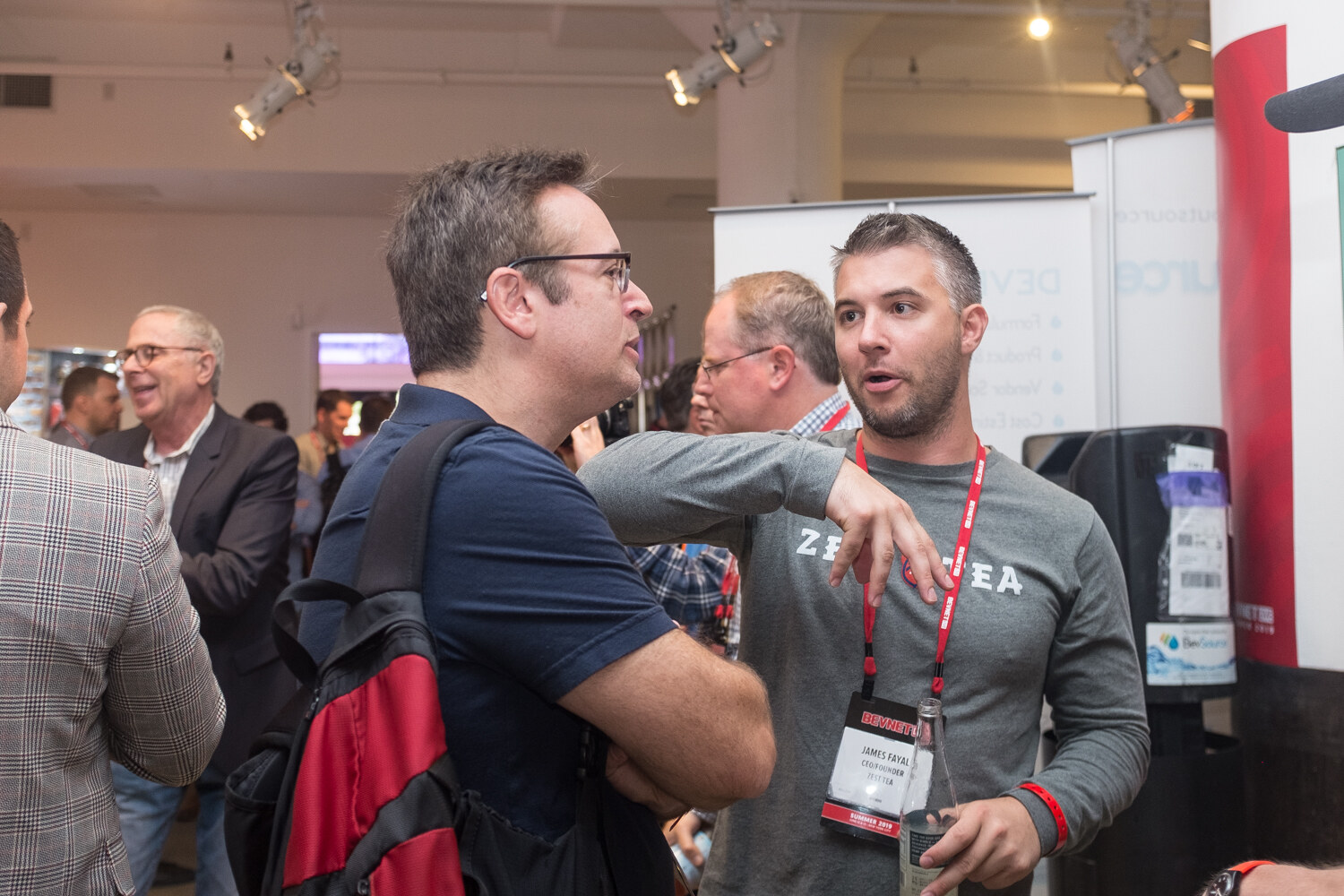 Man in a navy t-shirt and a man in a gray ZENA t-shirt talking at a booth.