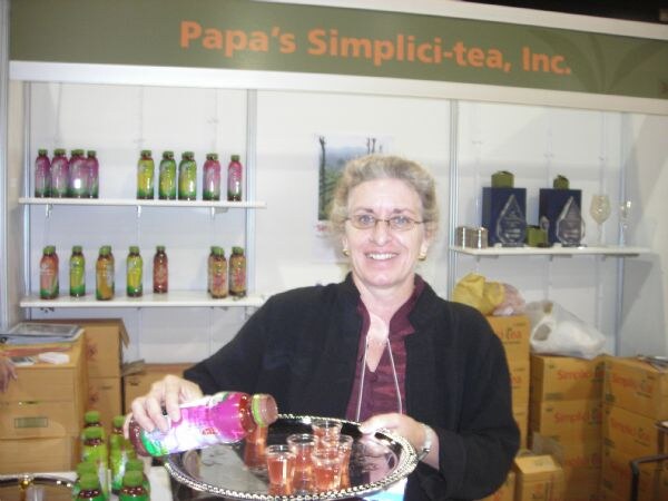 A smiling woman with glasses pours pink bottled tea into small cups on a silver tray at the Papa's Simplici-tea booth, with bottles on shelves and cartons behind her.