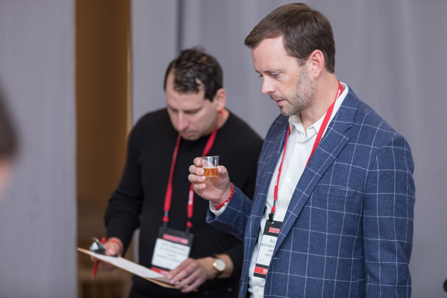 Two judges with red lanyards behind a gray curtain, one sipping a sample, before the semifinal.