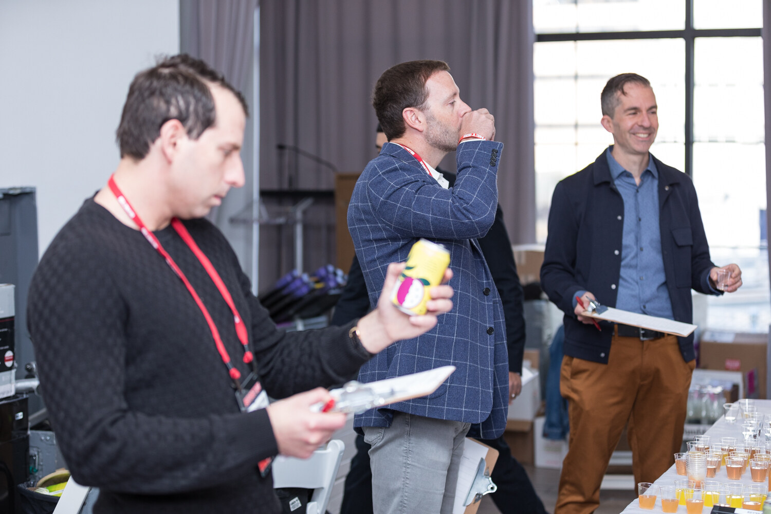 Judge in a black sweater holding a yellow can while others sample drinks near a window.
