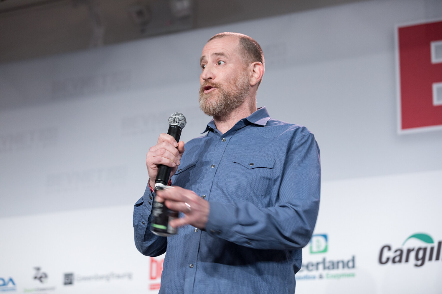 Bearded man in a blue button-up shirt speaking into a microphone in front of Berlin Packaging, Deerland and Cargill signs.