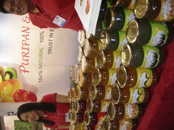 Sideways photo of many jars of fruit tea stacked on a red tablecloth at the Puripan booth, with two people in red shirts in the background.
