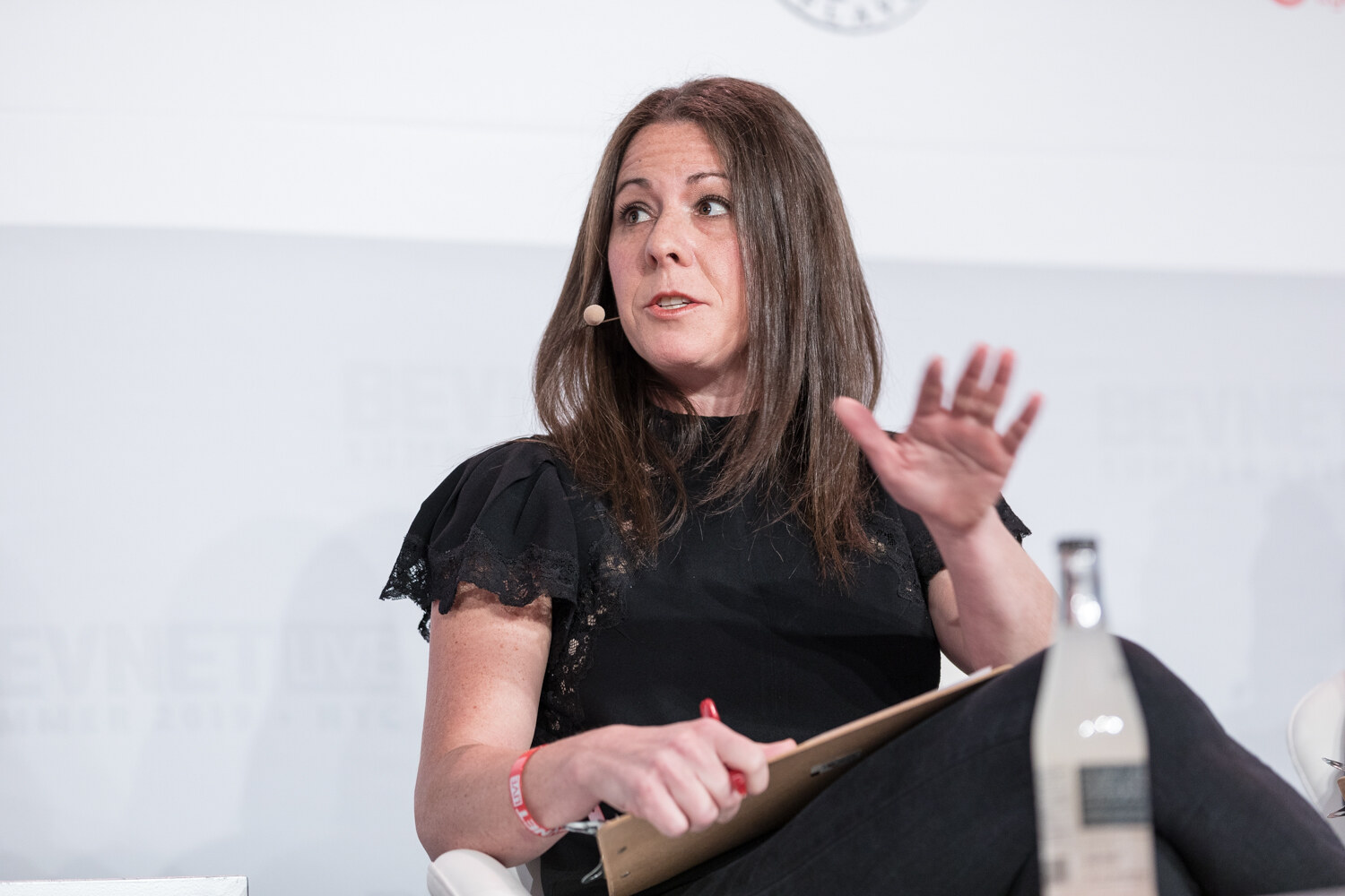 Judge in a black top with ruffled sleeves gesturing with a clipboard on her lap and a bottle on the table.