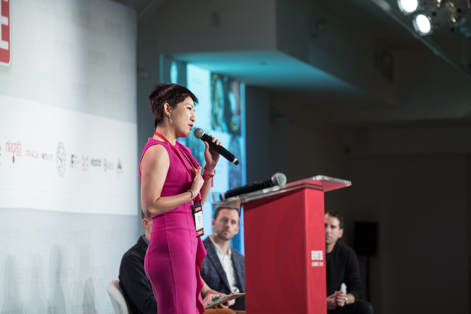 Woman in a magenta dress speaking into a microphone at the red podium, with judges seated behind her.