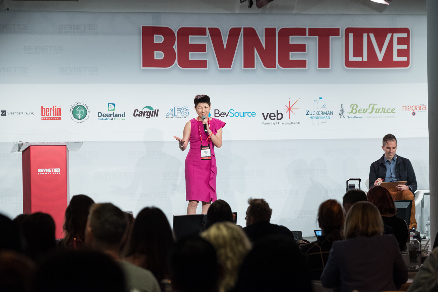 Wide view of a woman in a magenta dress on stage with the audience in the foreground and the BevNET Live logo above.