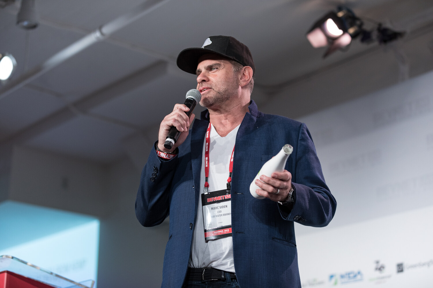 Man in a black cap and navy blazer holding a white bottle and microphone, with a lanyard around his neck.