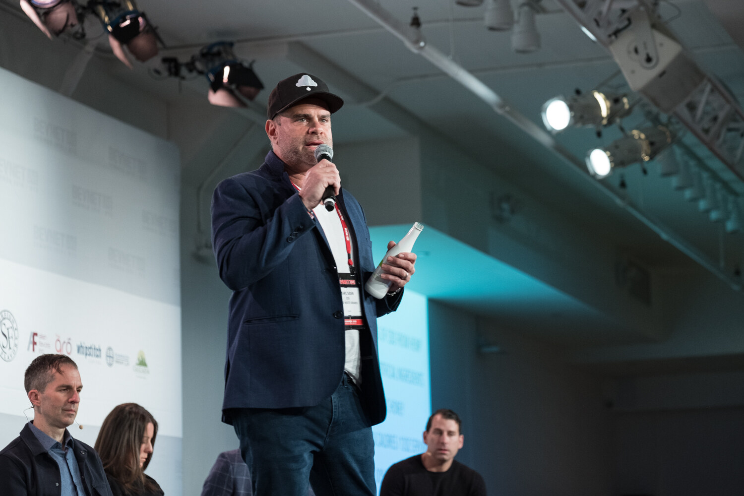Man in a black cap and navy blazer holding a white bottle and microphone, with judges seated behind him.
