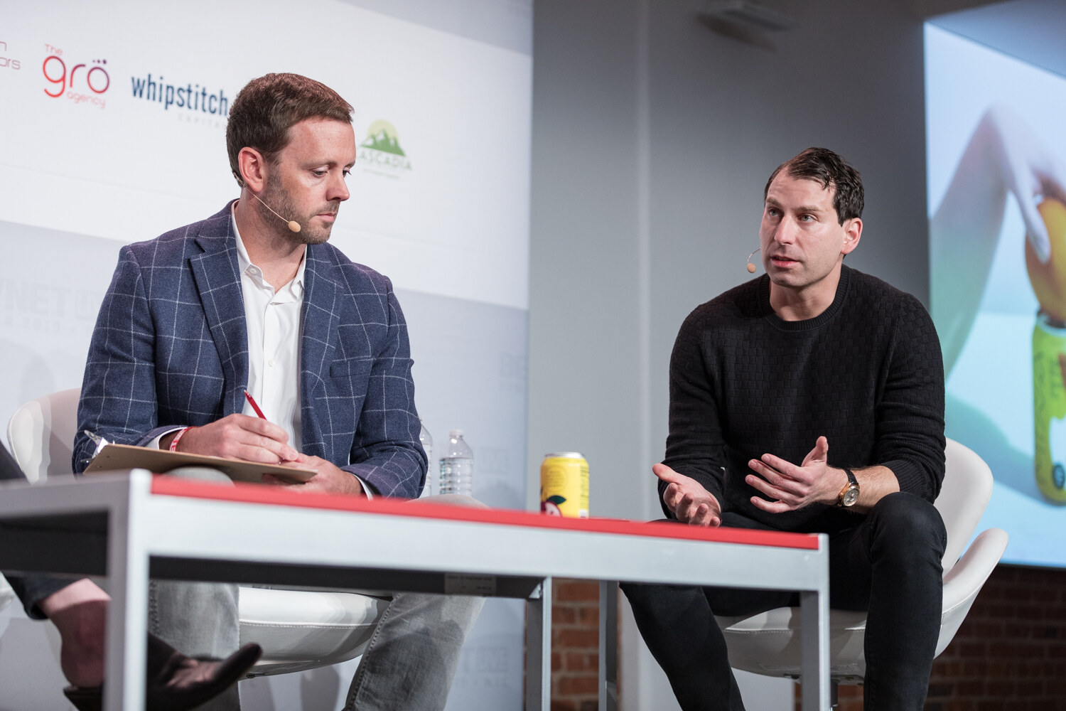 Two judges at the red table with a yellow can on it beside a slide showing a bottle.