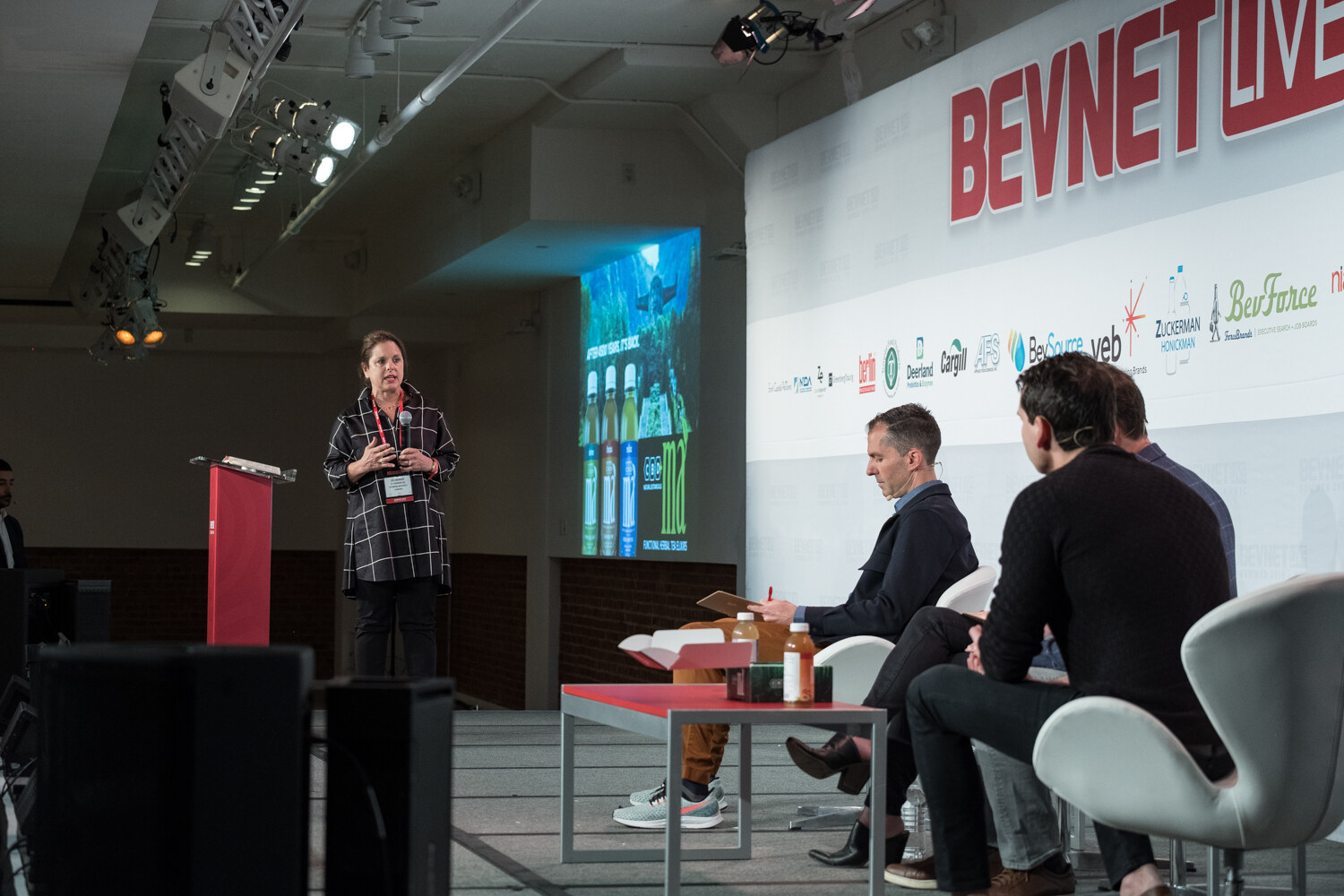 Woman in a black and white windowpane shirt speaking beside the red podium with a slide of bottles behind her.