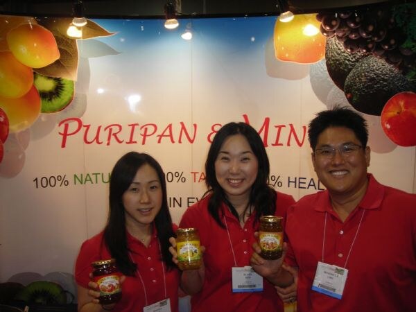 Two women and a man in red shirts stand smiling at the Puripan & Mind booth, holding jars of fruit tea in front of a backdrop with fruit images.