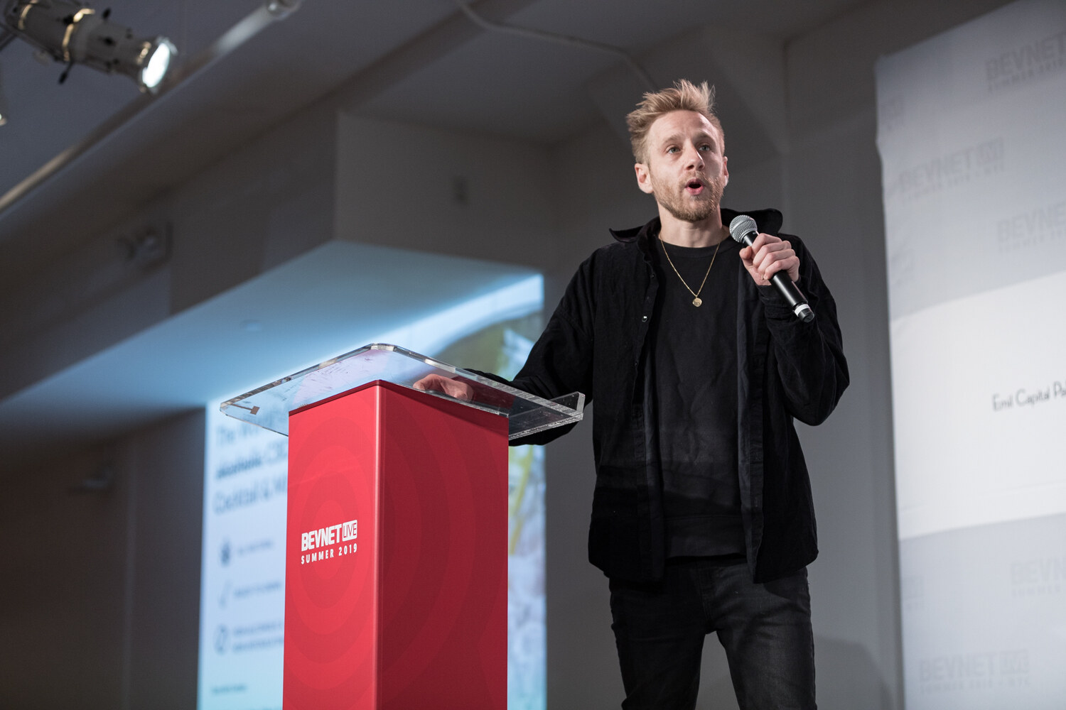 Man with spiky blond hair in a black overshirt standing beside the red podium, holding a microphone.