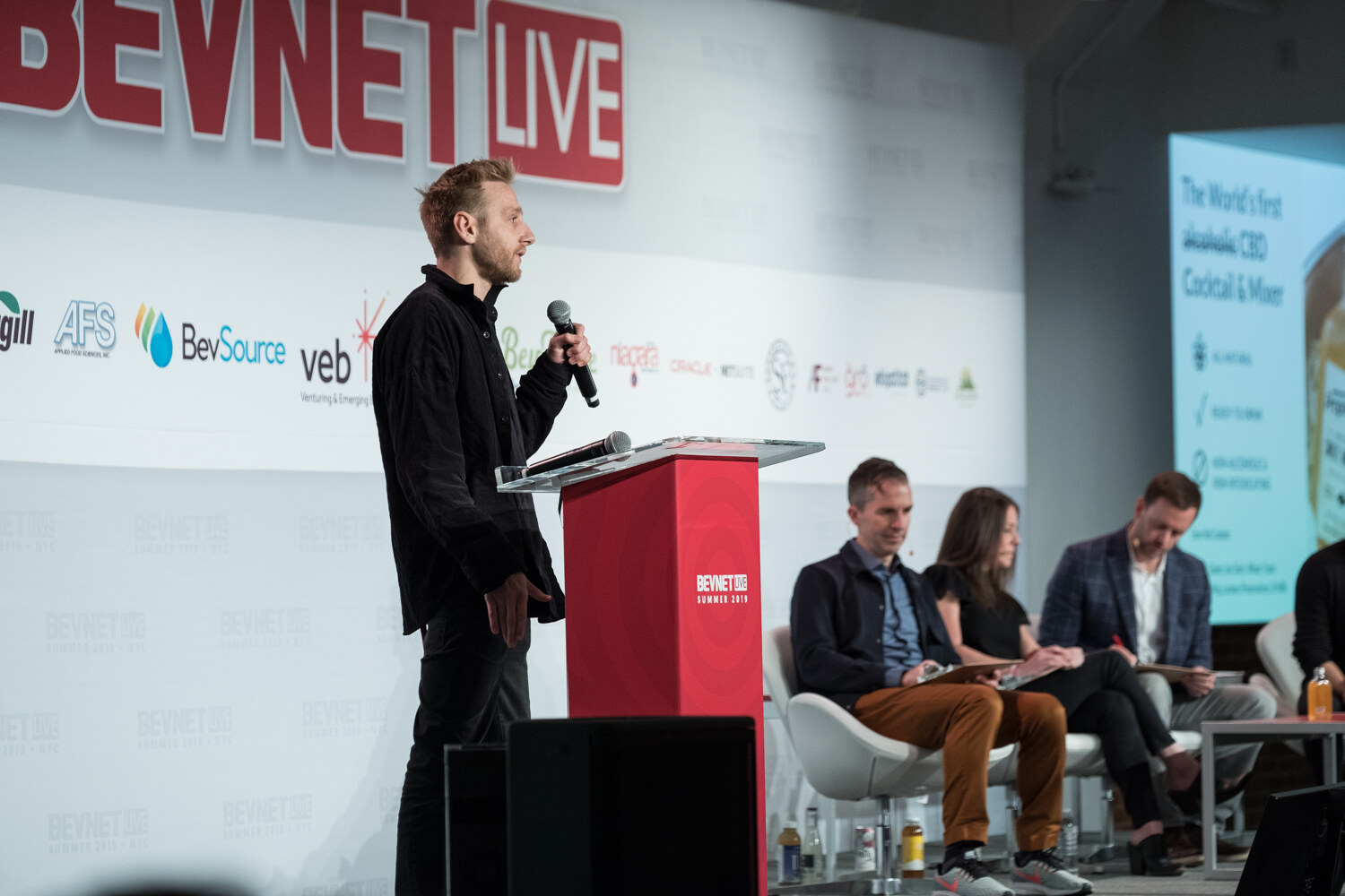 Man in black speaking at the red podium with a slide describing a first alcohol-free CBD cocktail and mixer, judges seated at right.