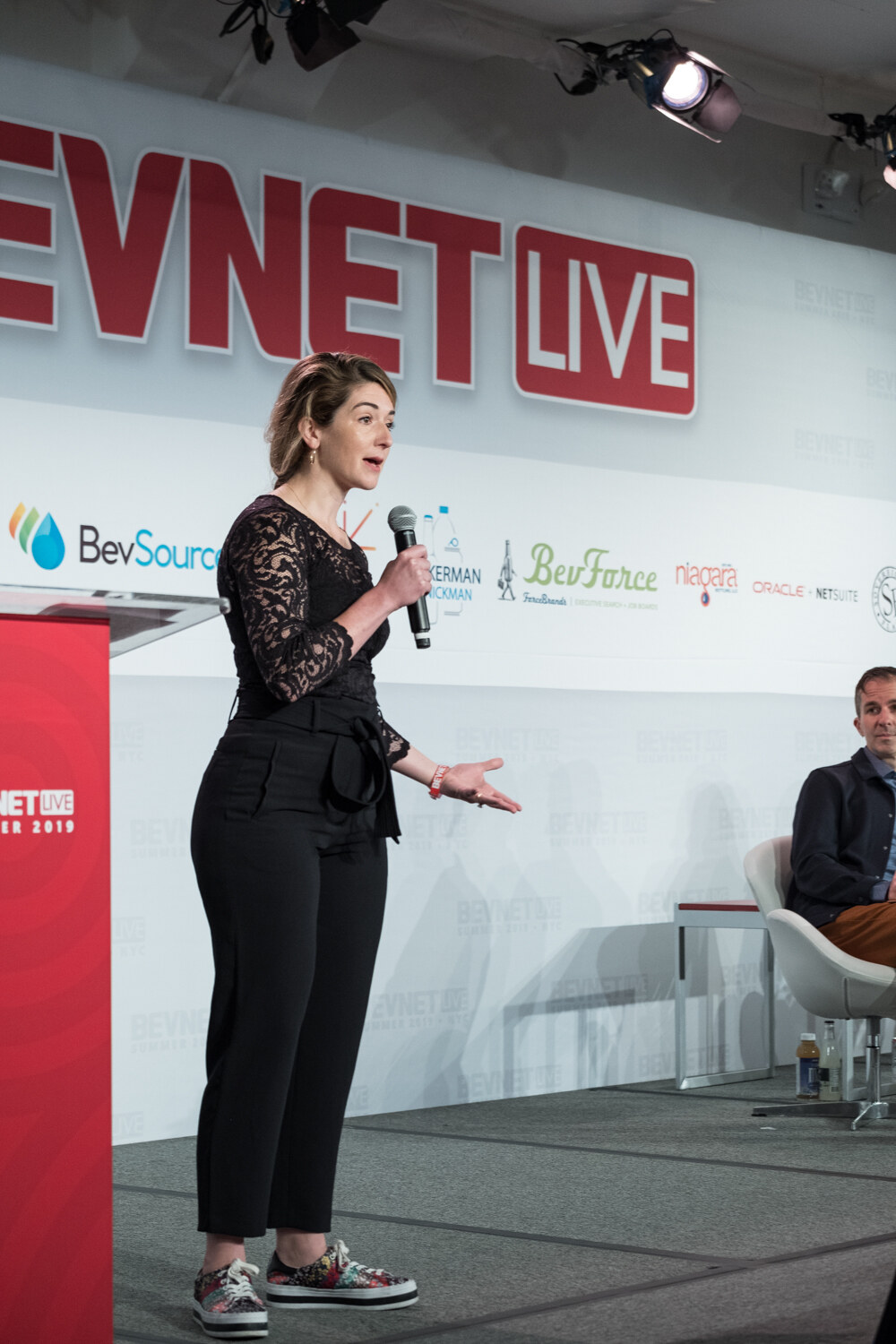 Woman in a black lace top and sneakers gesturing as she speaks in front of the BevNET Live sign.