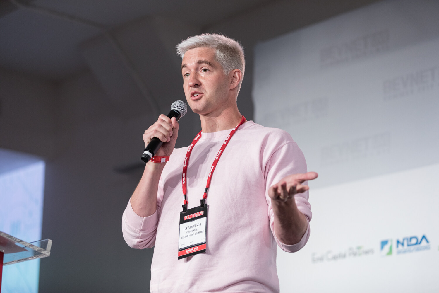 Presenter with silver-blond hair in a pink sweatshirt gesturing while speaking into a microphone.