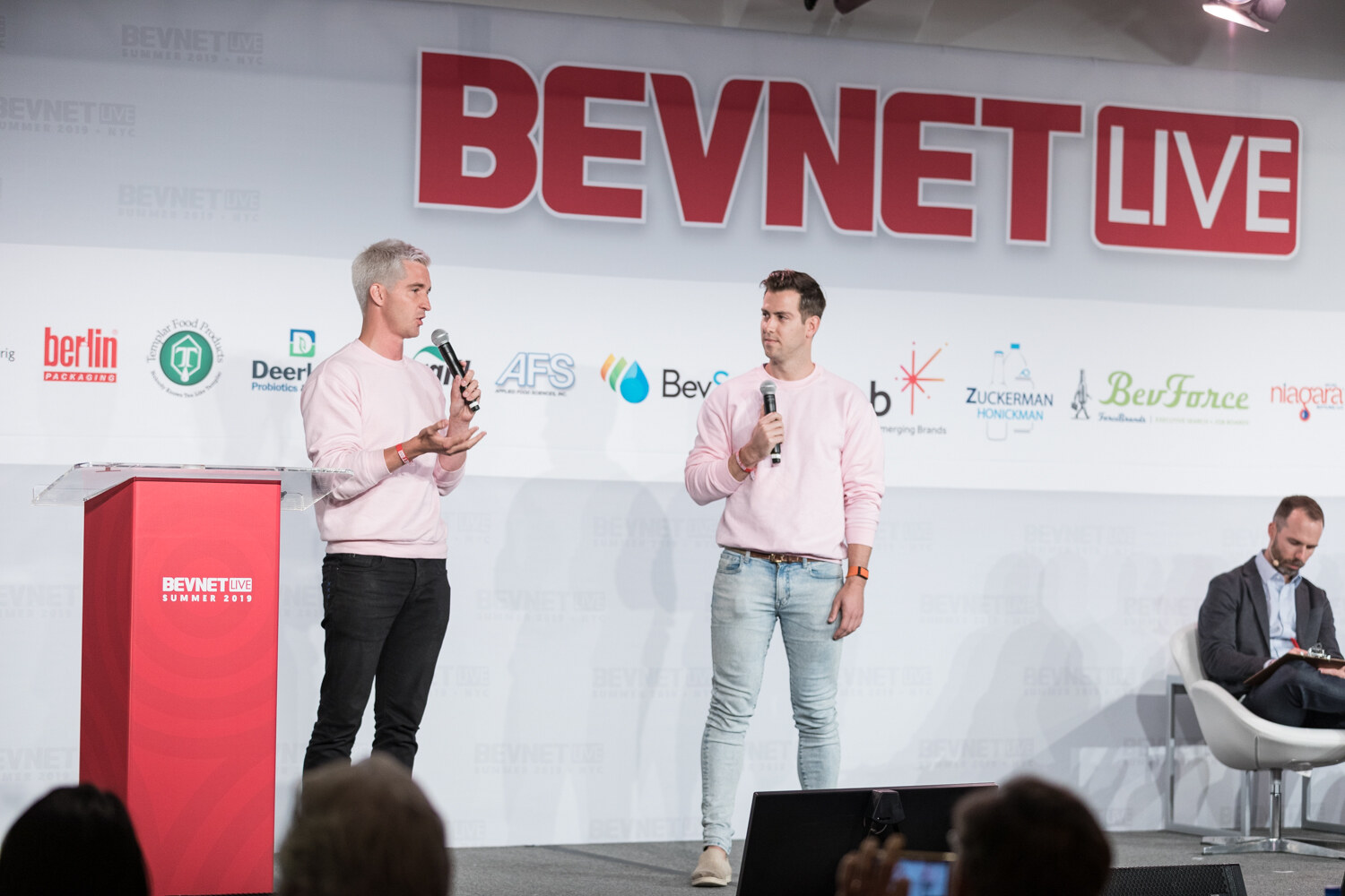 Two men in pink sweatshirts standing on stage beside the red podium, with a judge visible at right.