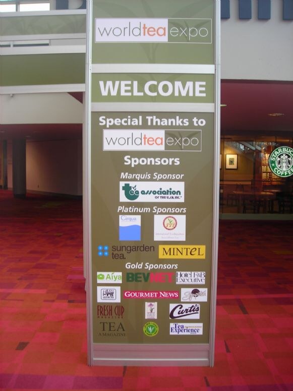 A World Tea Expo welcome sign lists marquis, platinum and gold sponsors with logos, standing on red patterned carpet in front of a Starbucks Coffee shop.