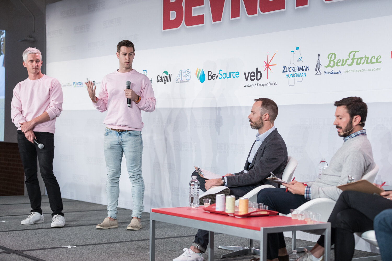 Two men in pink sweatshirts presenting on stage while the judges sit at a table lined with sample cups.